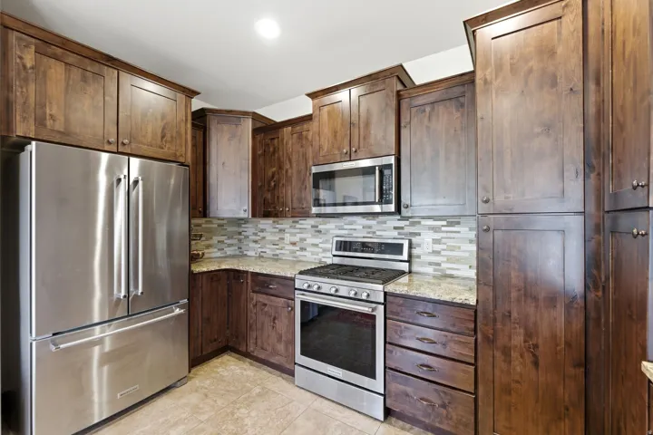 Kitchen featuring stainless steel appliances, wood-finish cabinetry, granite countertops, tile flooring, and a mosaic tile backsplash