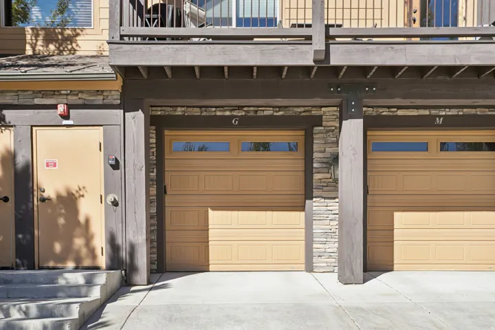 Two beige garage doors with horizontal window panels, flanked by stone veneer accents and dark wood structural beams