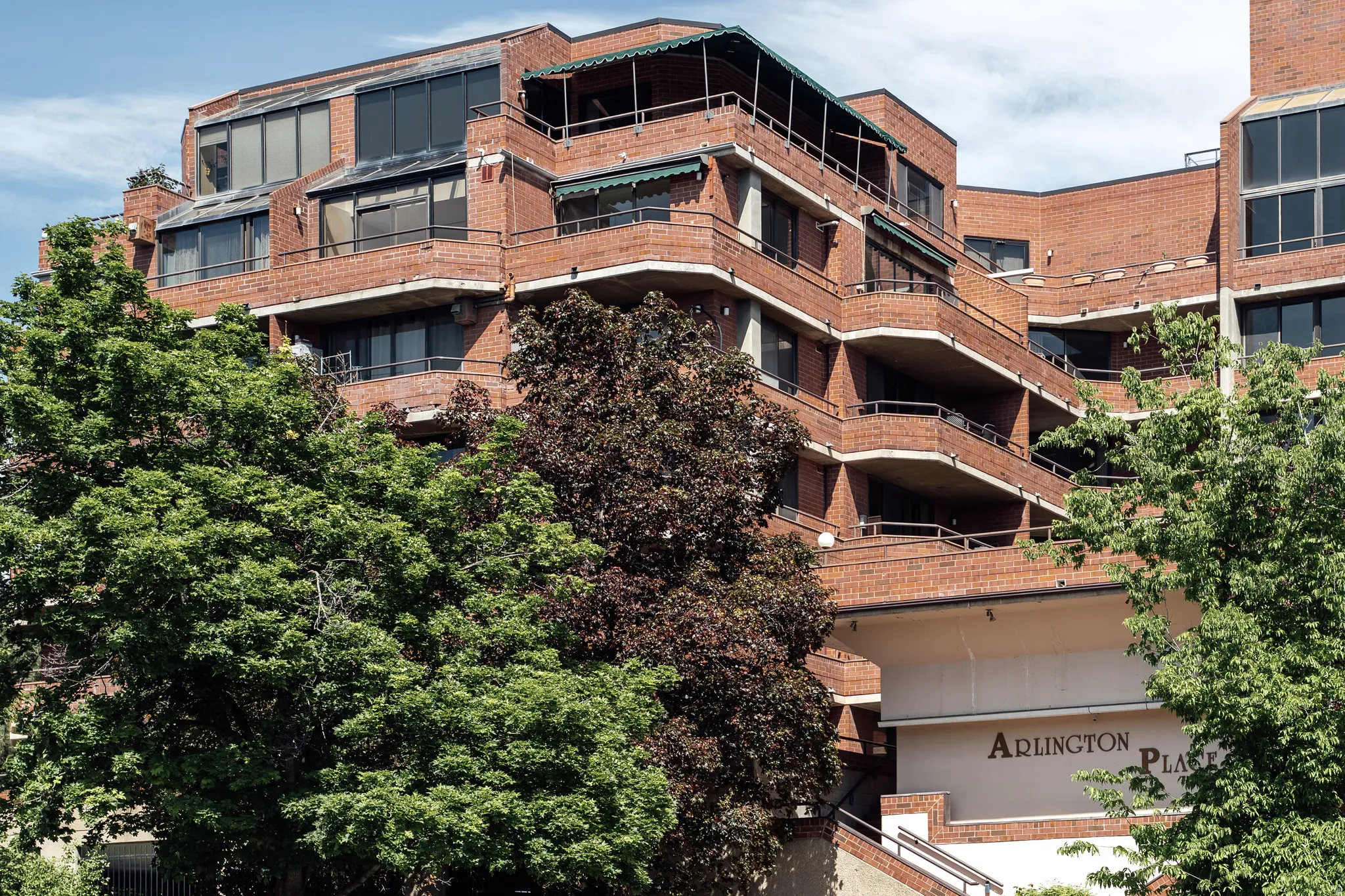 Brick multi-story building featuring multiple balconies with metal railings, a distinct architectural design with varied setbacks, large windows, and a prominent "Arlington Place" sign