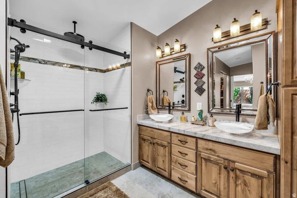 Dual vanity with light wood cabinetry and vessel sinks, complemented by a large walk-in shower featuring a sliding glass door and subway tile surround