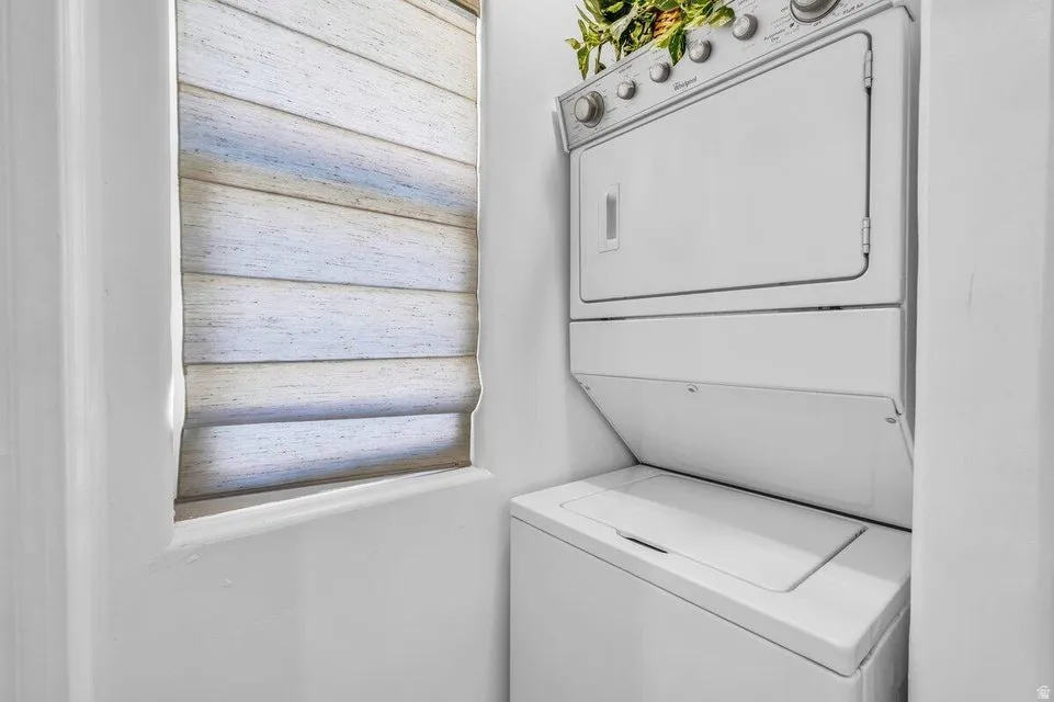 White stacked washer and dryer unit with a nearby window featuring horizontal blinds