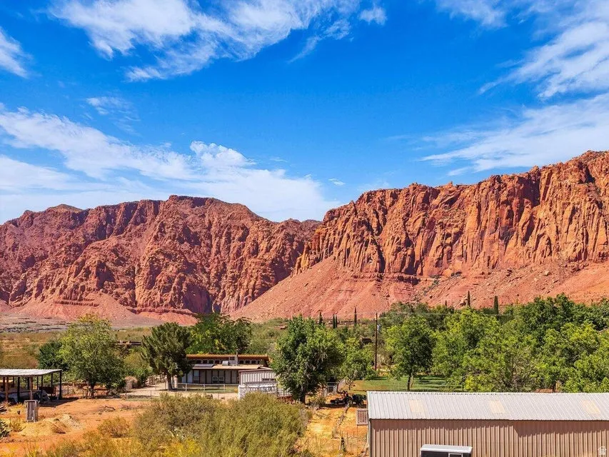 Expansive landscape featuring significant red rock formations, a clear blue sky with scattered clouds, and a property with multiple structures