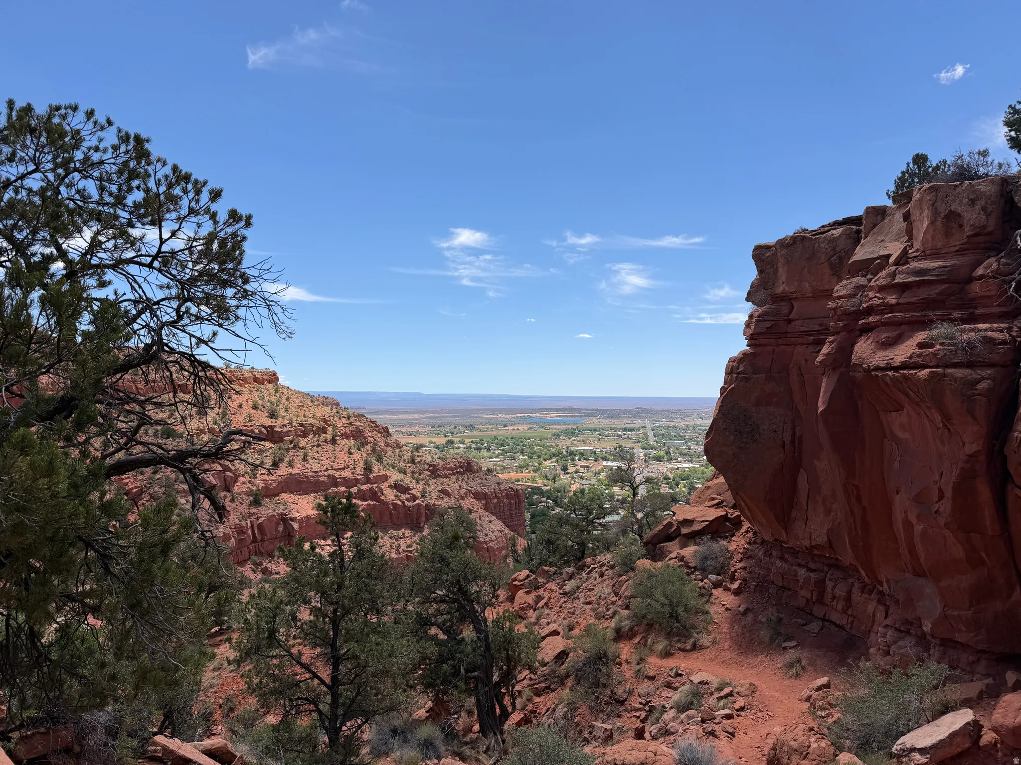 Expansive red rock landscape featuring distant town below, vibrant blue sky, scattered green foliage, and natural rock formations