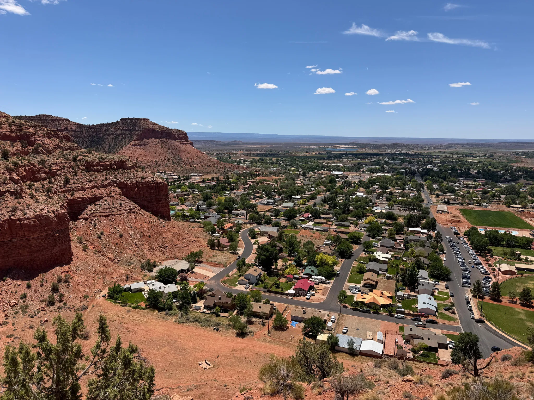 Expansive aerial capturing a community featuring residential properties, mature green landscaping, and paved roadways, set against a backdrop of natural red rock formations and distant open terrain