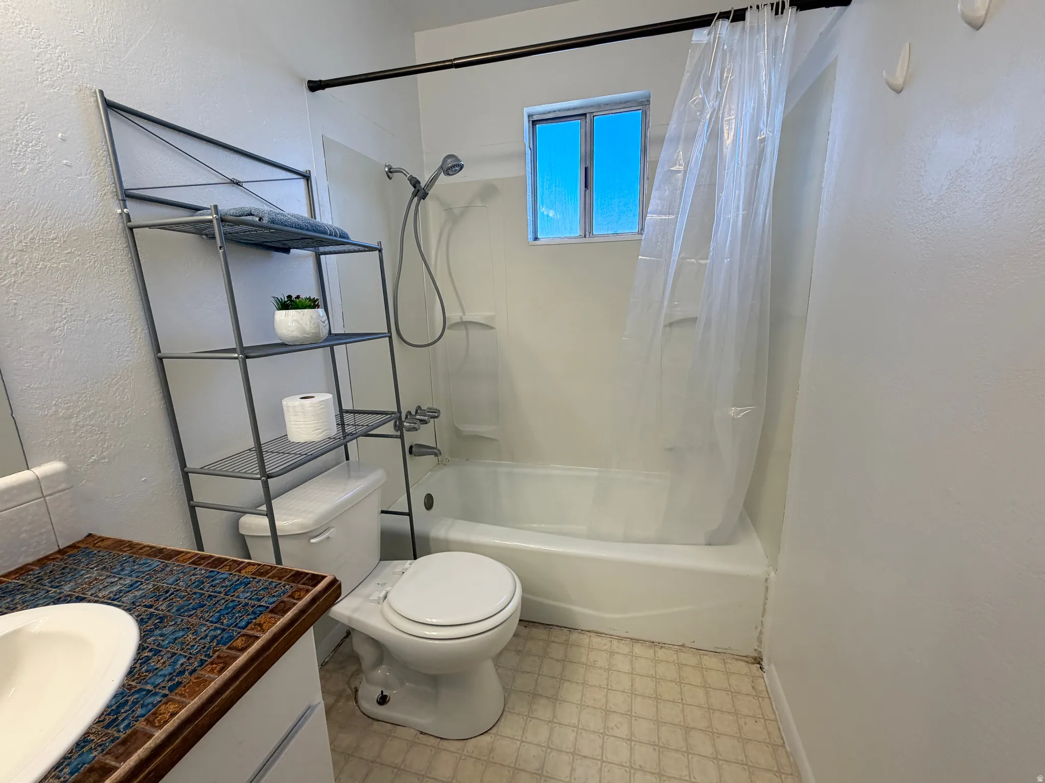 Bathroom featuring a white bathtub with an integrated shower, a window, and a vanity with a patterned tile countertop and integrated sink