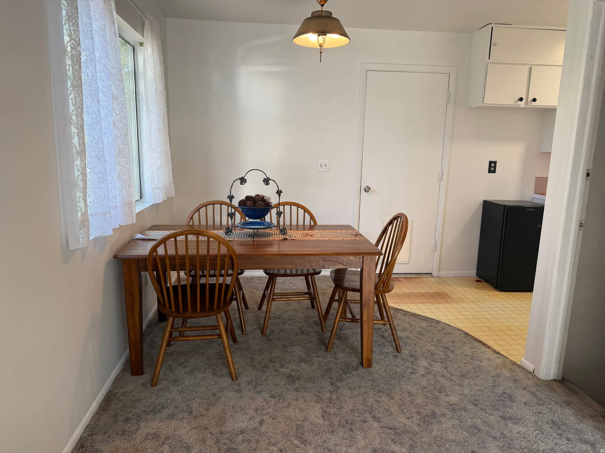 Dining area featuring light gray carpeting, a window with sheer curtains, a white door, overhead light fixture, and white upper cabinetry