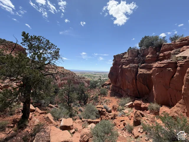 Red rock landscape featuring prominent cliff formations, scattered green foliage, and a distant valley under a clear blue sky