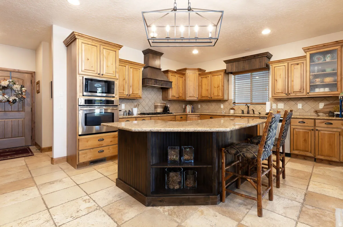 Spacious kitchen featuring an angled island with a granite countertop, light wood cabinetry, stainless steel double ovens, and tile flooring