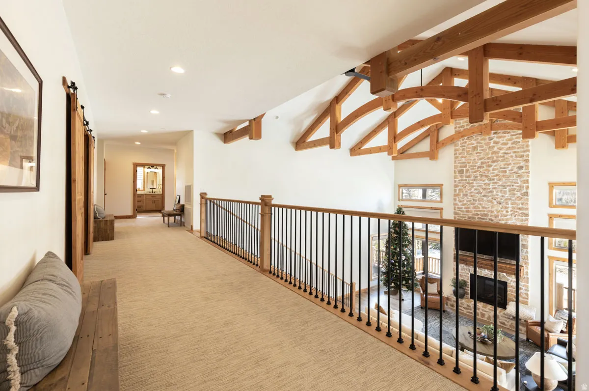 Upper-level hallway overlooking a great room featuring vaulted ceilings with exposed wood beams, a stone fireplace, and a light wood railing with black spindles