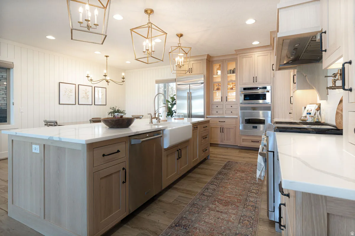 Spacious kitchen featuring an expansive center island with an integrated farmhouse sink, light wood-finish cabinetry, stainless steel appliances, and wood-finish flooring