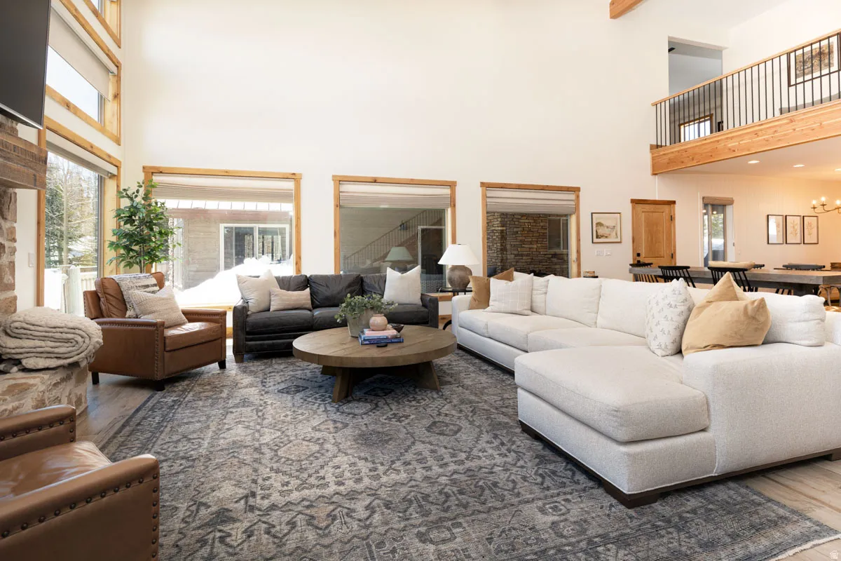 Spacious living area featuring double-height ceilings, large windows with natural wood trim, wood-finish flooring, a stone-clad fireplace, and an open-concept layout with a visible second-story overlook
