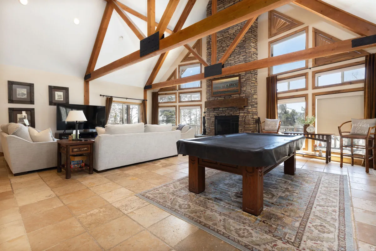 Vaulted ceilings with exposed wood beams, stone-faced fireplace with wood mantel, large windows with natural wood trim, and tile flooring