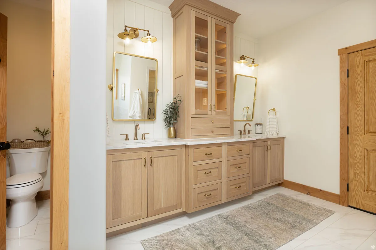 Dual vanity with light wood-finish cabinetry, white countertops, and brushed brass hardware