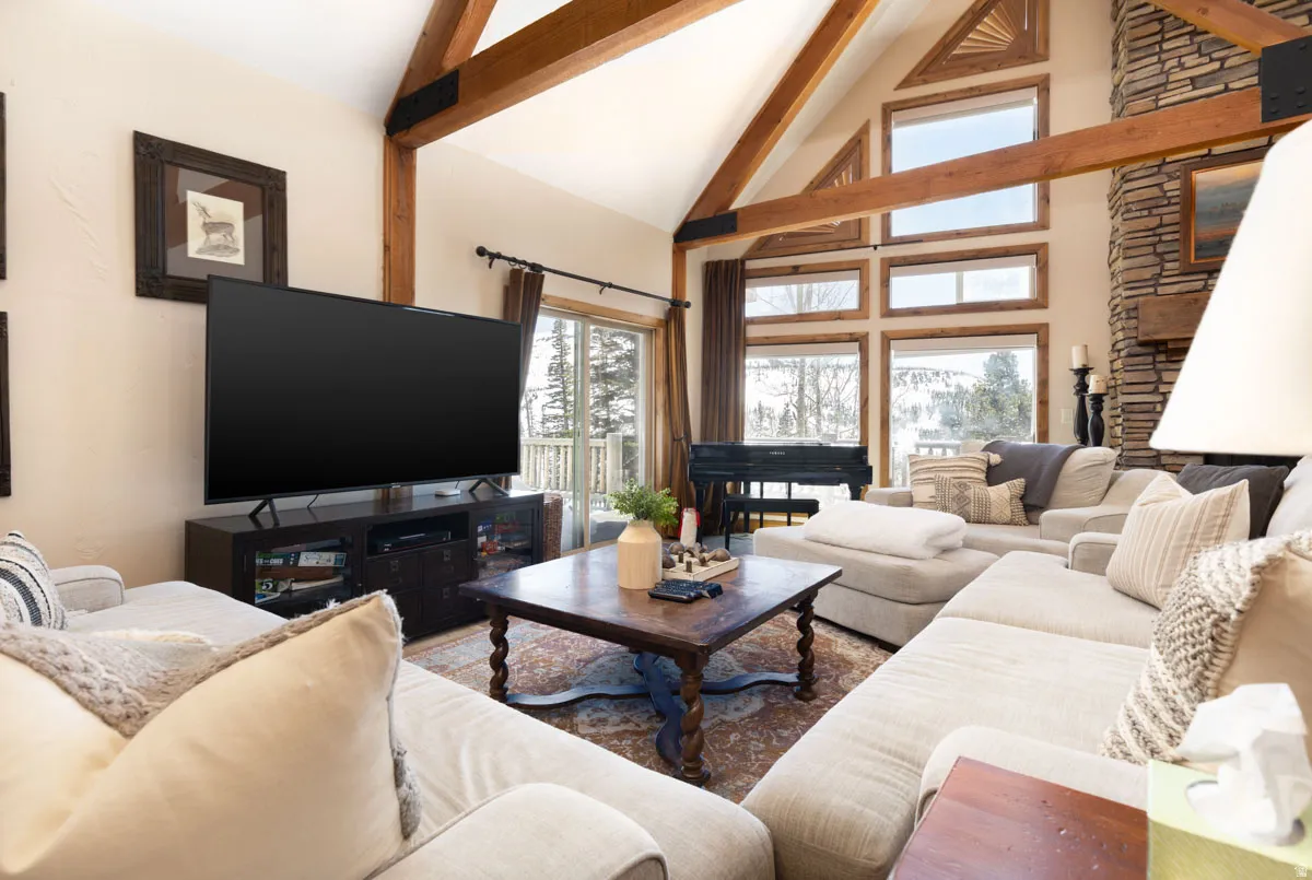 Living area featuring vaulted ceilings with exposed wood beams, a stone fireplace, large picture windows, and a sliding glass door leading to a deck