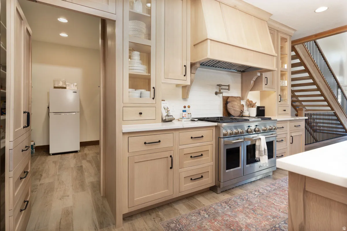Gourmet kitchen featuring a professional-grade range, custom wood cabinetry, a built-in wood range hood, light-toned countertops, and a full-height tile backsplash