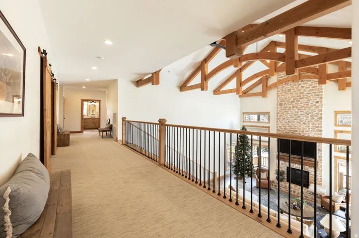 Upper-level hallway overlooking a great room featuring vaulted ceilings with exposed wood beams, a stone fireplace, and a light wood railing with black spindles