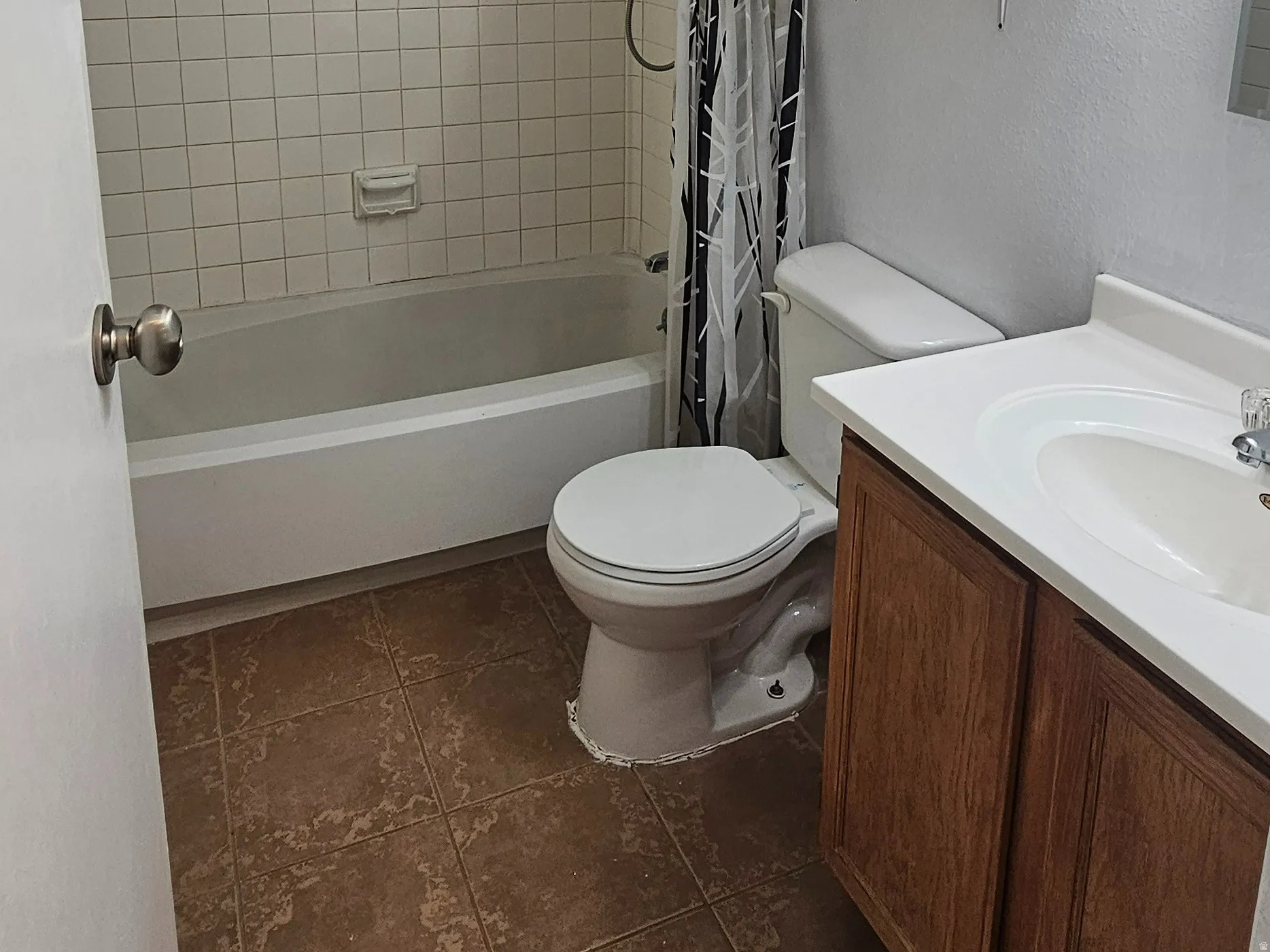 Bathroom featuring a white tub with tile surround, a wood-finish vanity with a white countertop and integrated sink, and brown tile flooring
