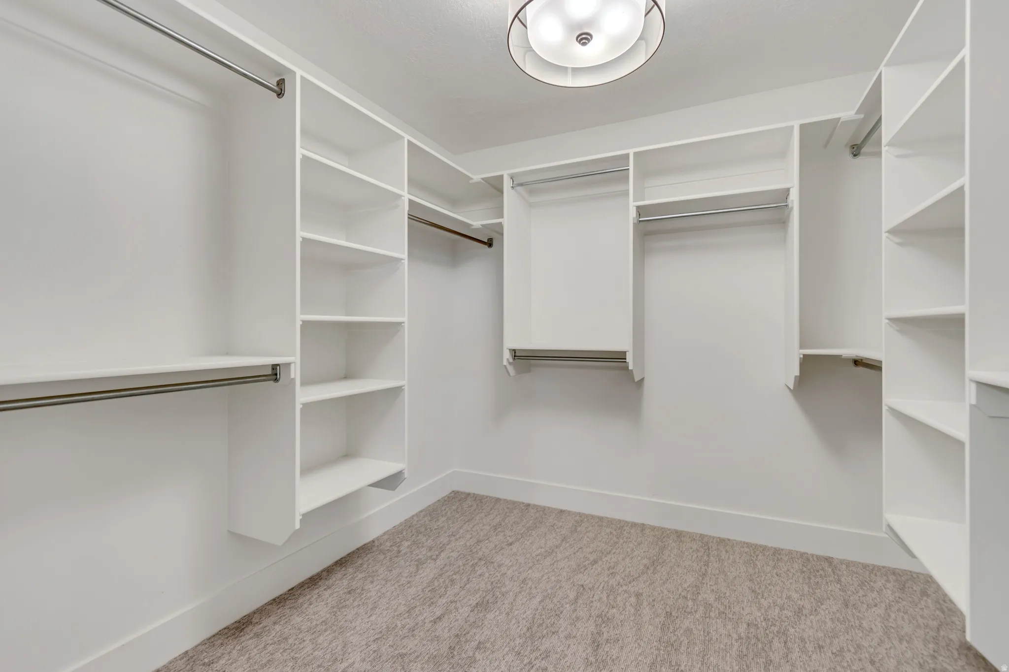 Spacious walk-in closet featuring extensive built-in shelving, multiple hanging rods, clean white finishes, light neutral-toned carpeting, and a modern flush-mount light fixture
