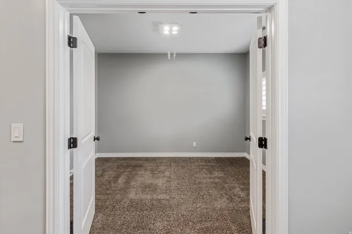 Double door entry into a room featuring light gray walls, plush carpeting, white baseboards, and a recessed ceiling light fixture
