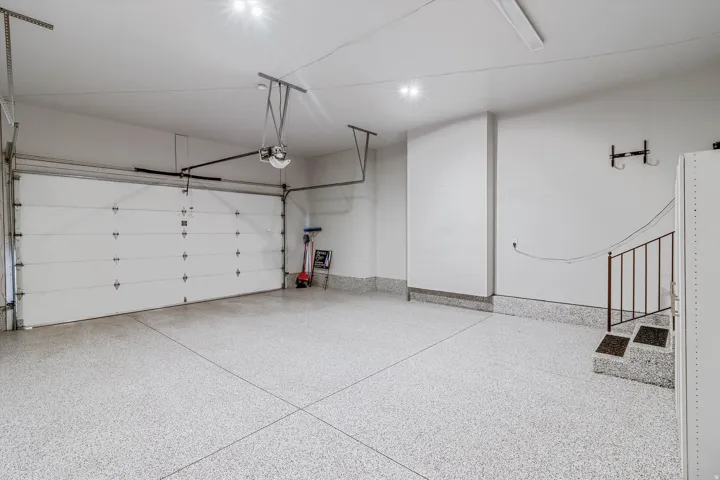 Spacious garage featuring a speckled epoxy floor, white paneled garage door, high ceilings, recessed lighting, and a built-in staircase with metal railing
