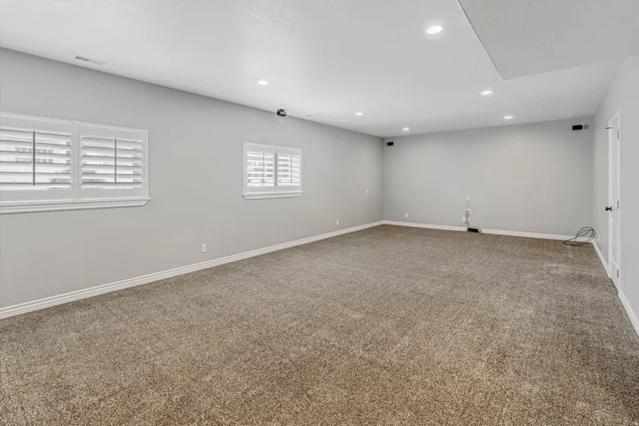 Spacious room featuring neutral carpeting, light gray walls, two windows with white plantation shutters, recessed ceiling lighting, and a white interior door