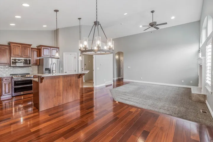 Open concept living space featuring rich wood-finish flooring in the kitchen, a spacious island with a light-toned countertop, stainless steel appliances, a tile backsplash, and a living area with high vaulted ceilings and a fireplace with a light-toned m