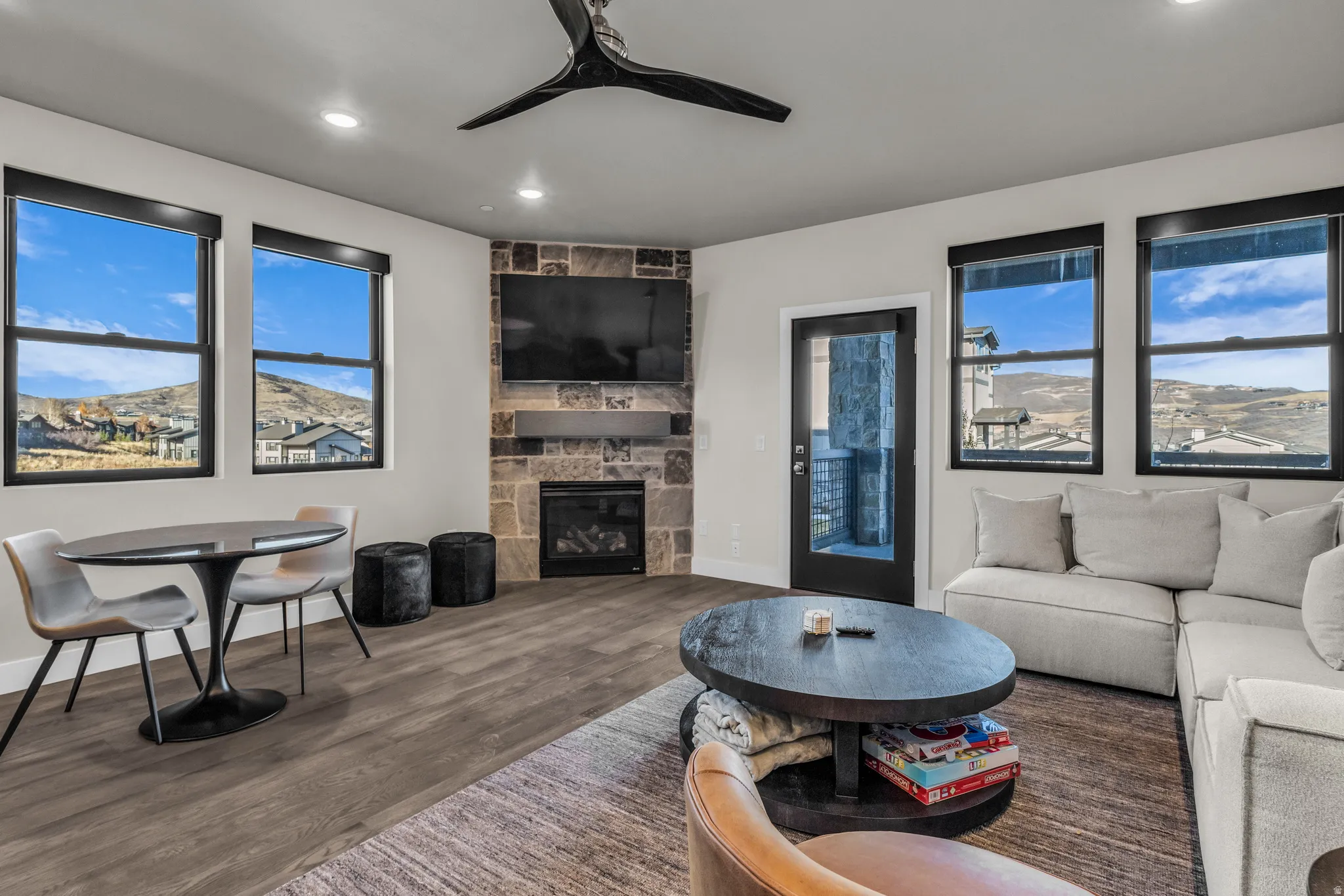 Living area featuring wood-finish flooring, a stone-clad fireplace with a natural wood mantel, recessed lighting, and multiple windows with black frames