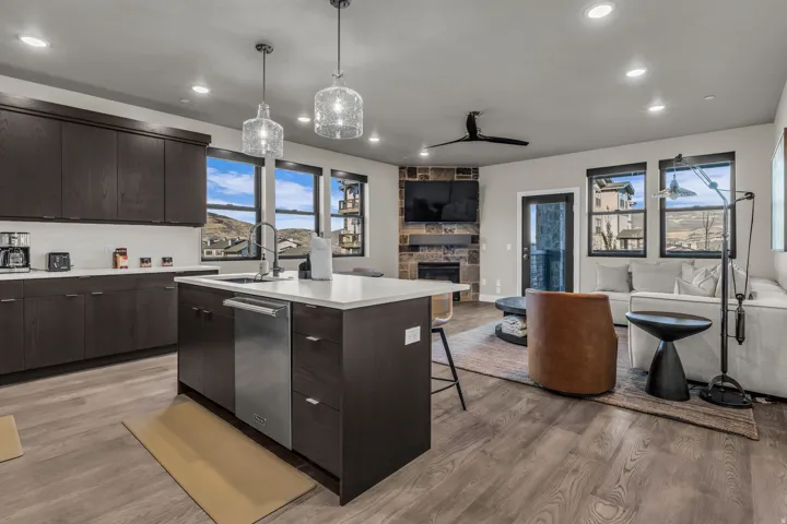 Modern kitchen and living area featuring dark wood-finish cabinetry, a central island with a white countertop and integrated stainless steel dishwasher, and wood-finish flooring