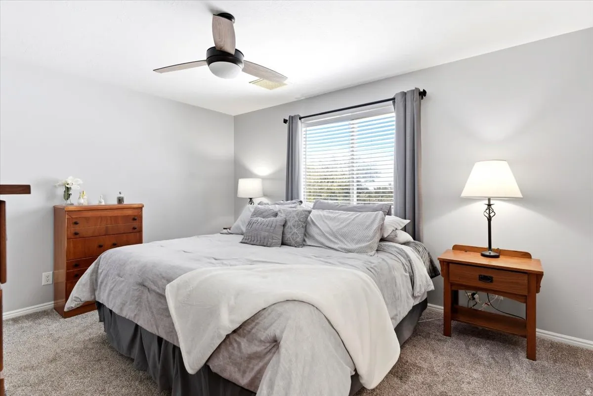 Carpeted interior space featuring light gray wall paint, a ceiling fan with integrated lighting, and a window with blinds and gray curtains