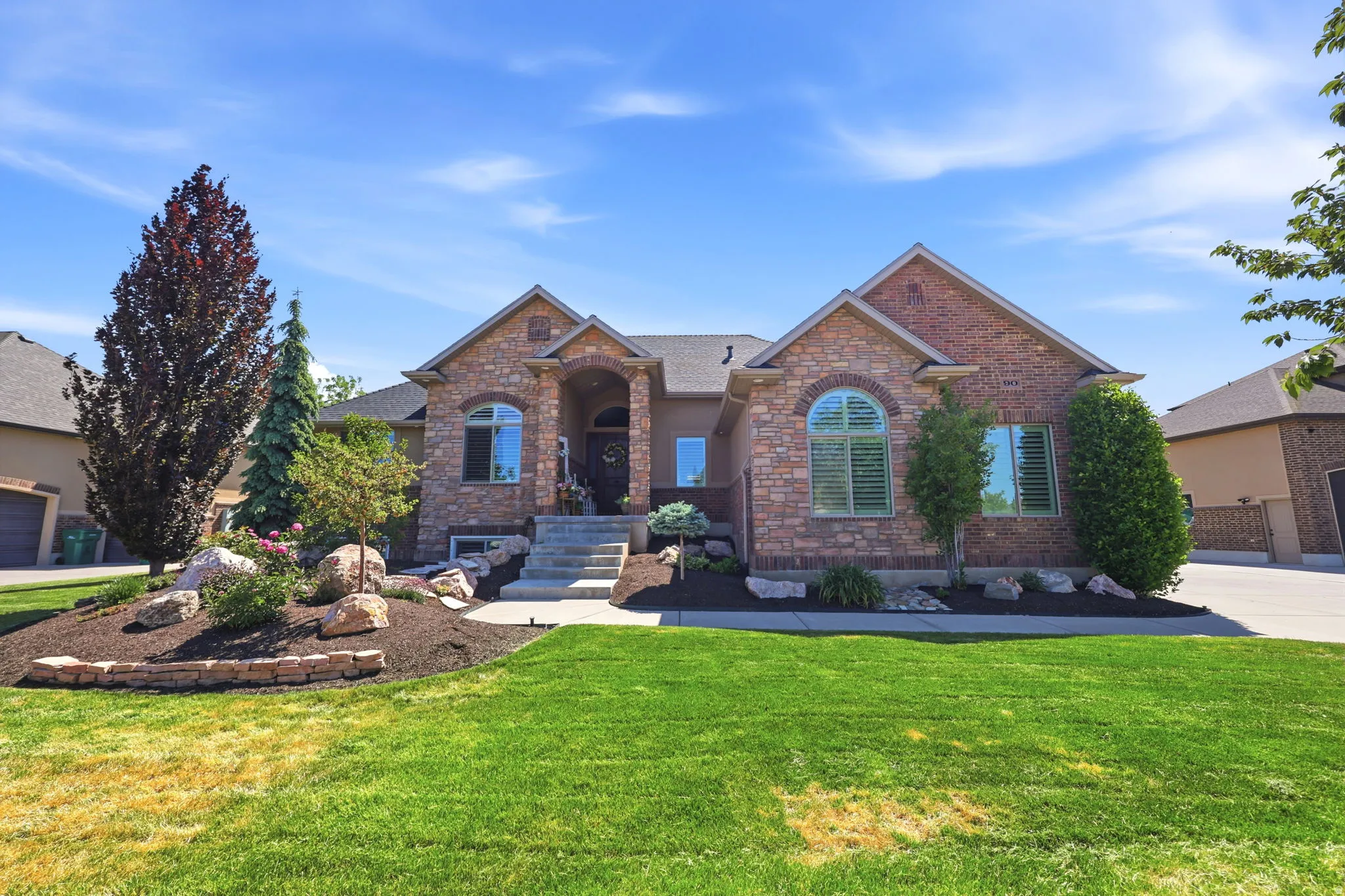 Stone and brick facade residence with an arched entry and covered porch, featuring mature landscaping, a sloped front lawn, and an integrated two-car garage
