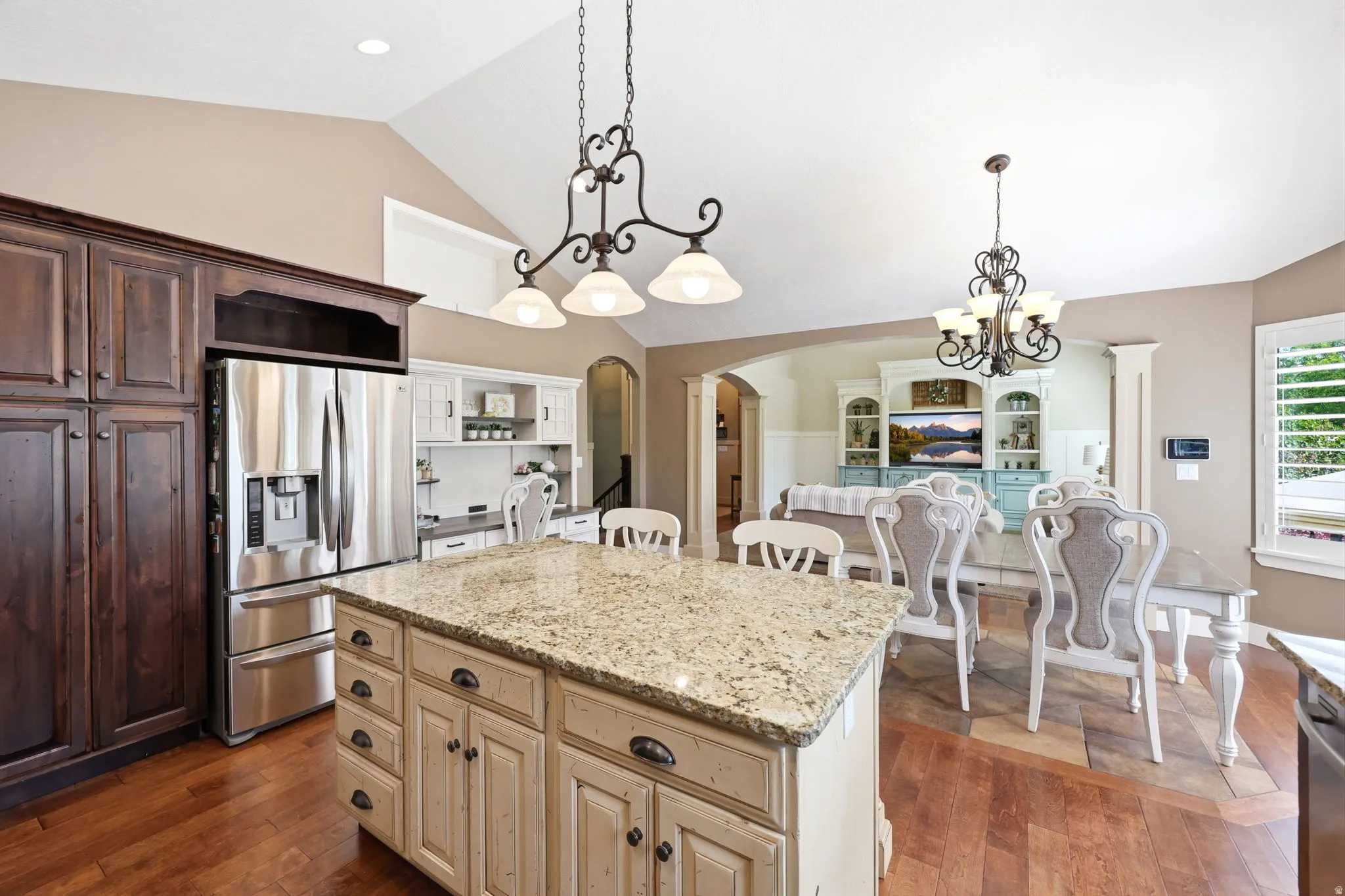 Kitchen featuring a central island with granite countertop, wood-finish flooring, dark wood cabinetry, a stainless steel refrigerator, and vaulted ceilings with decorative lighting fixtures