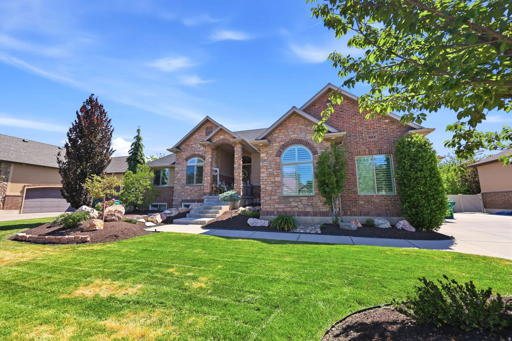 Brick and stone facade residence featuring a covered entry, arched window with plantation shutters, mature landscaping with rock accents, and a wide concrete driveway