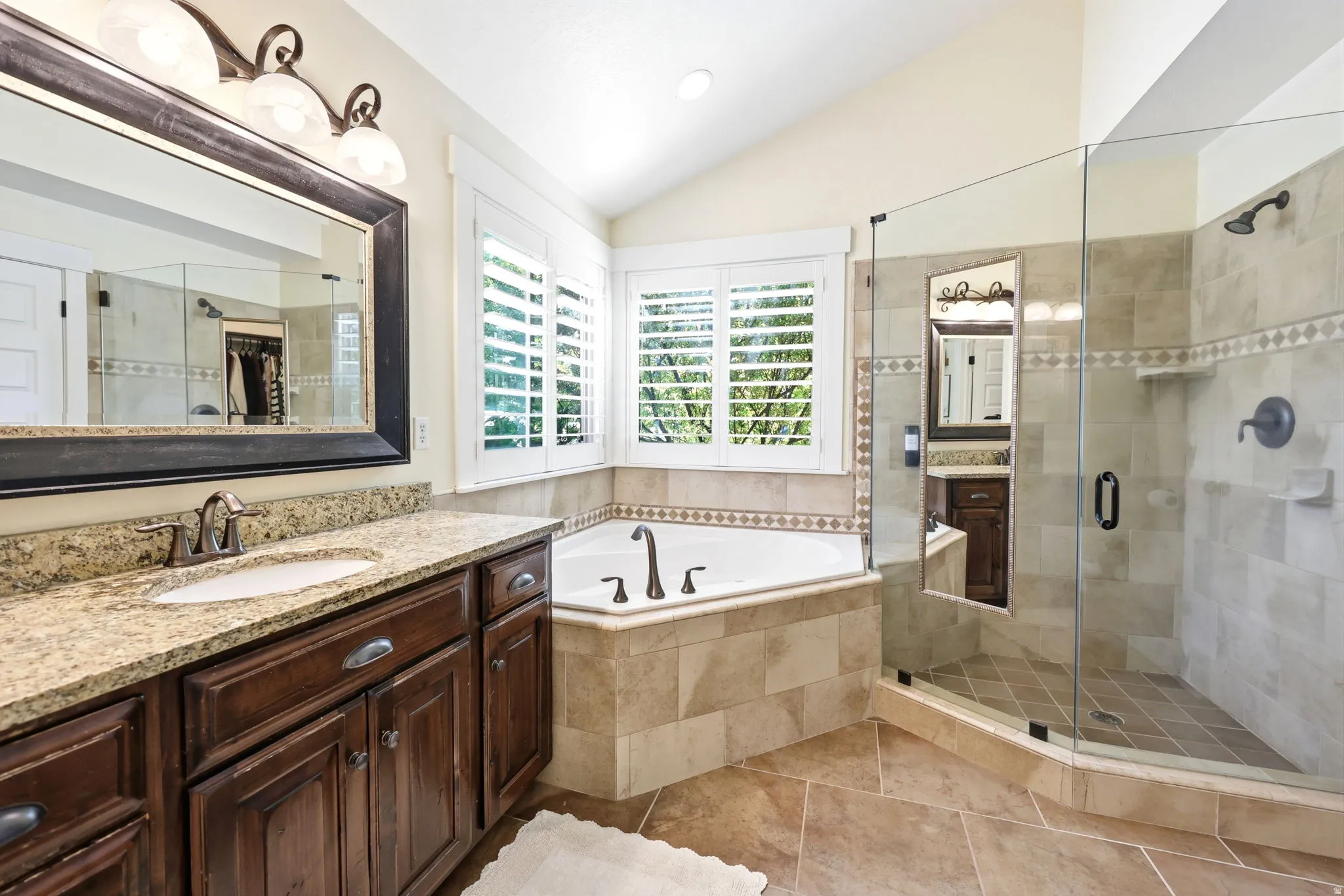 Spacious bathroom featuring a corner soaking tub, glass-enclosed shower with tile surround, wood vanity with a stone countertop, and plantation shutters