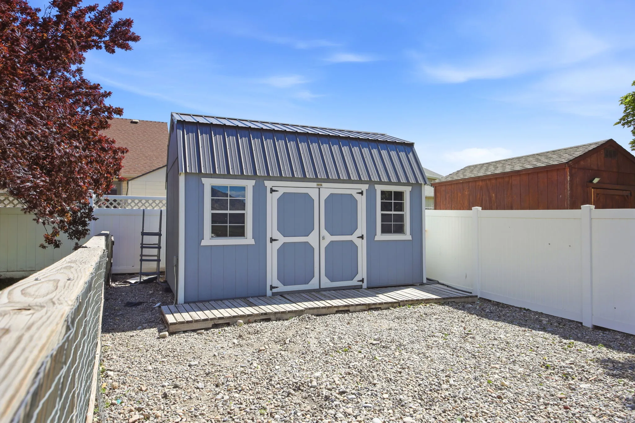 Blue gable-roofed shed with white trim and a metal roof, situated on a gravel pad