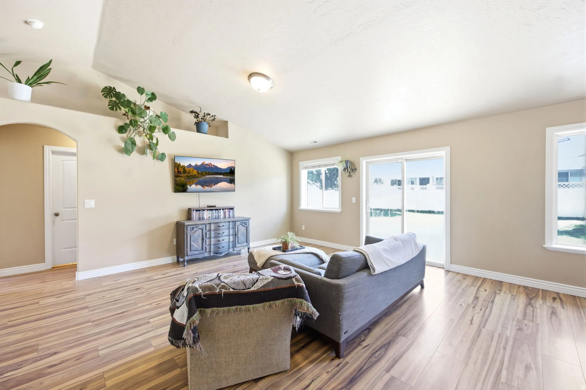 Spacious living area featuring vaulted ceilings, wood-finish flooring, an arched doorway, a sliding glass door, and multiple windows providing natural illumination
