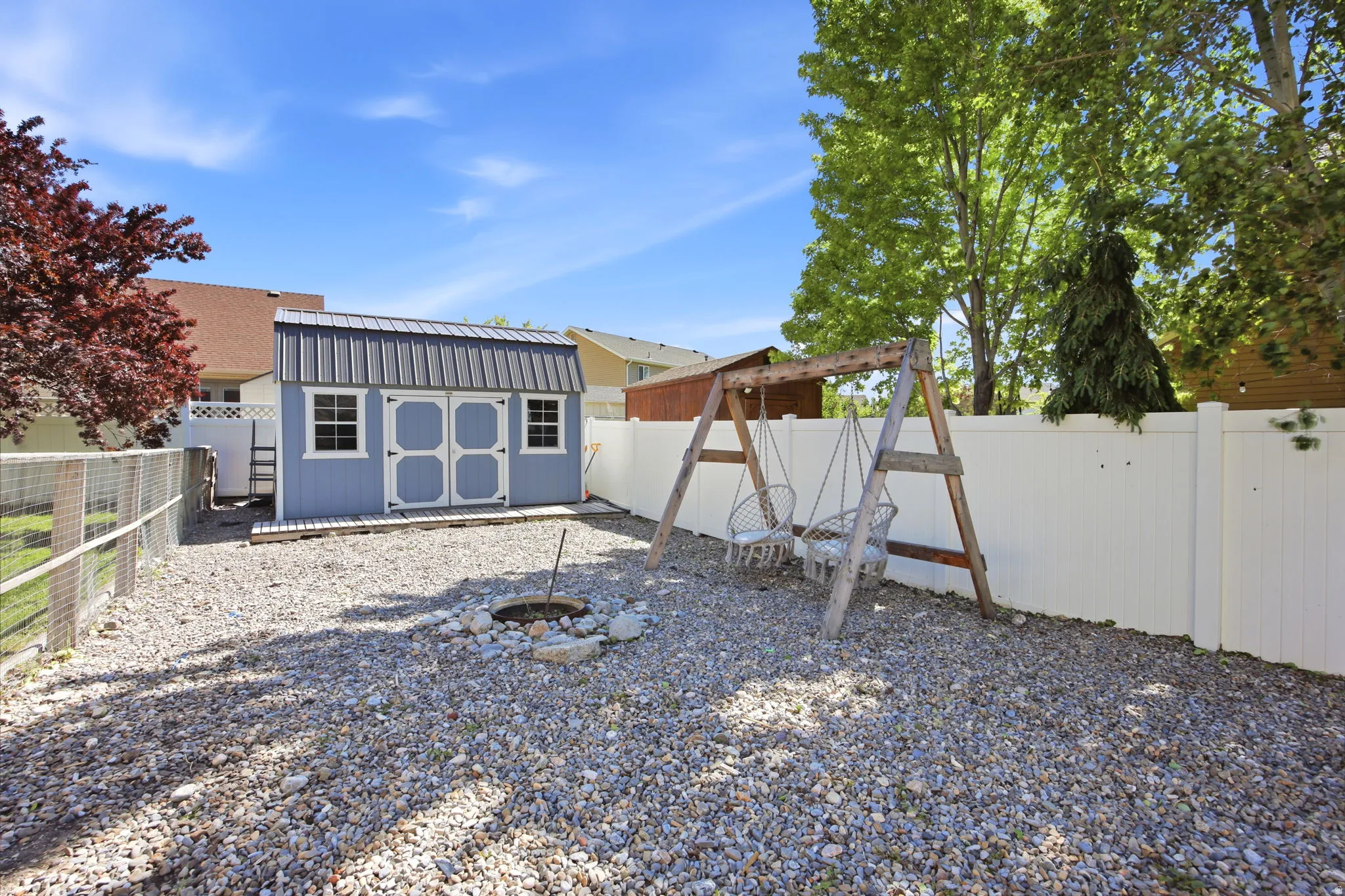 Spacious gravel yard featuring a blue storage shed with a metal roof, a white privacy fence, and a built-in fire pit