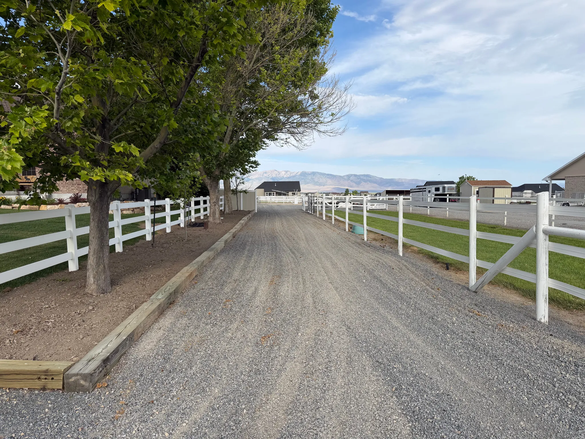 Gravel drive featuring white post-and-rail fencing, mature trees, and a mountain backdrop