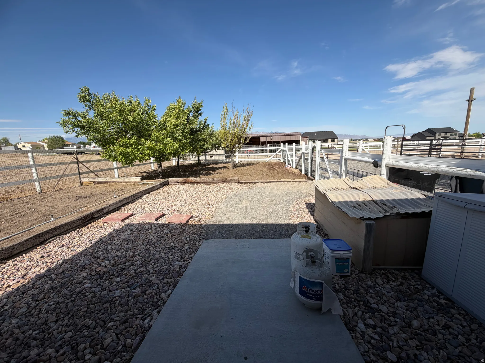 Gravel-finished outdoor area featuring mature trees, a white fence line, and a concrete patio section
