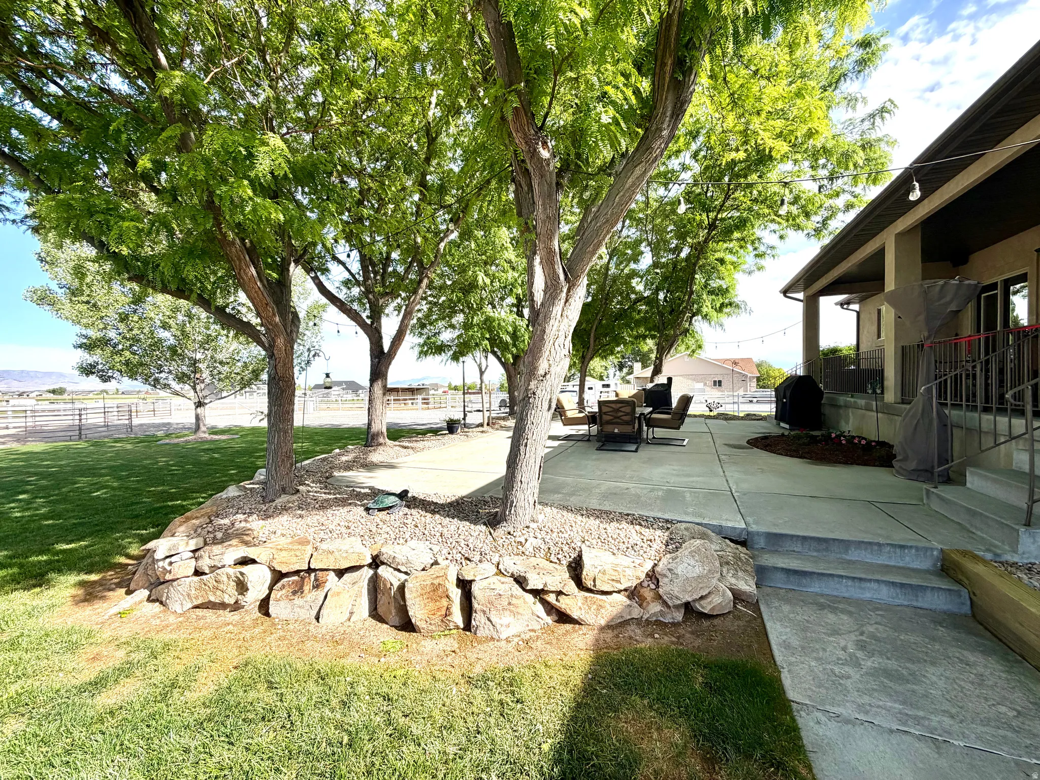 Expansive concrete patio area shaded by mature trees, featuring a stone retaining wall and terraced steps