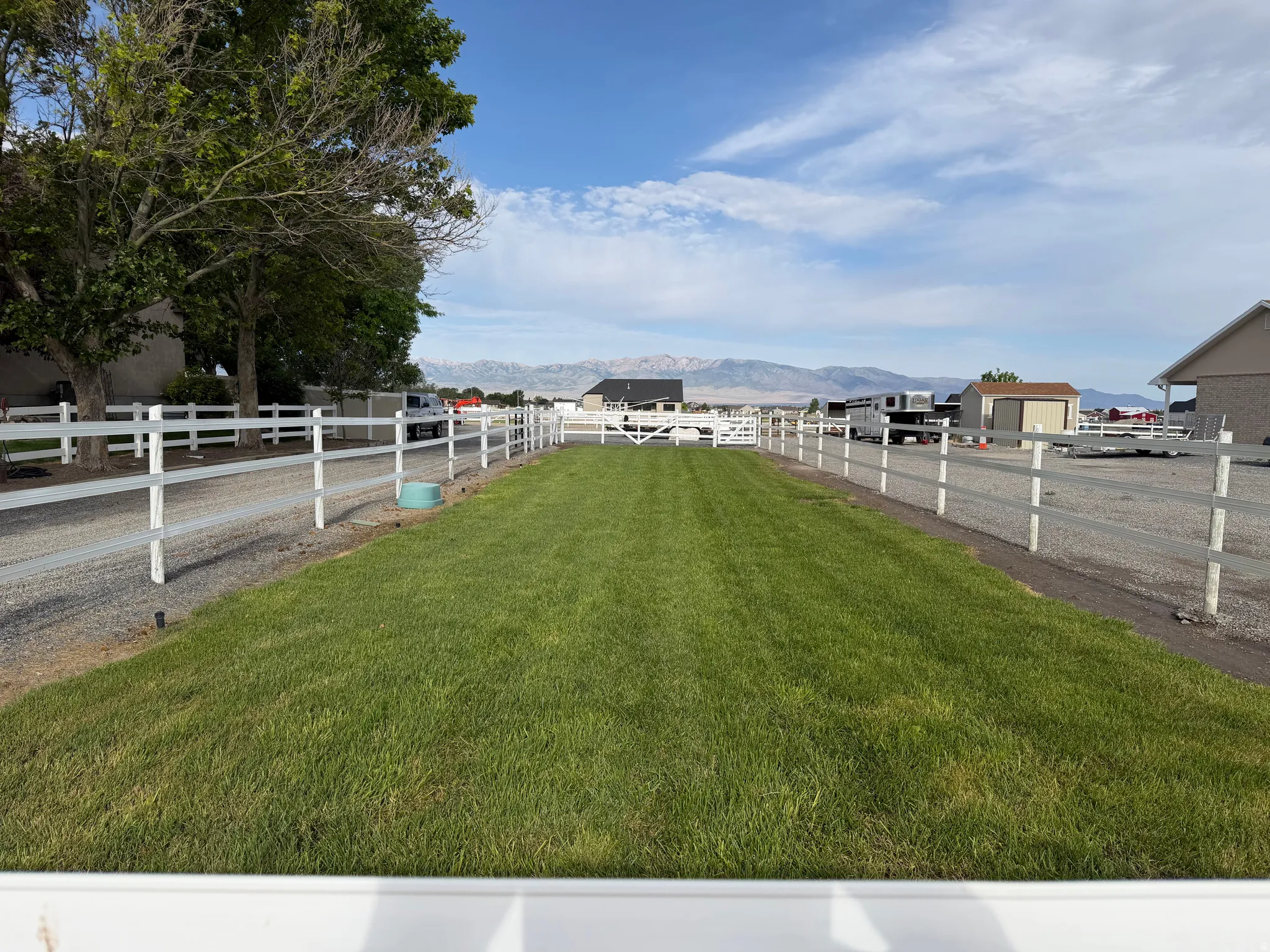 Expansive lawn area featuring white vinyl fencing, a gravel driveway, and distant mountain scenery
