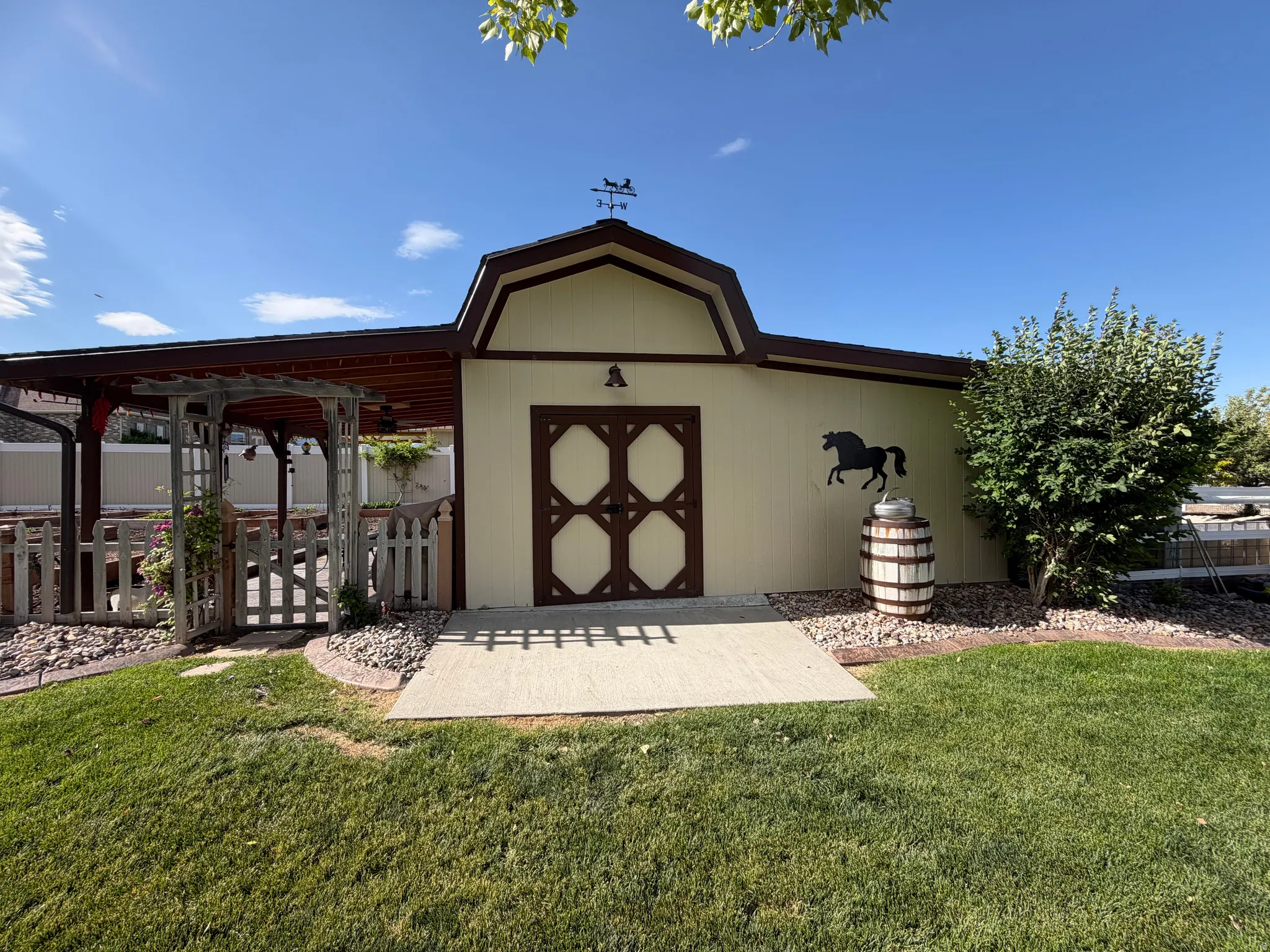 Cream-colored outbuilding with a gambrel roofline, featuring decorative doors, a weather vane, and an attached wood pergola