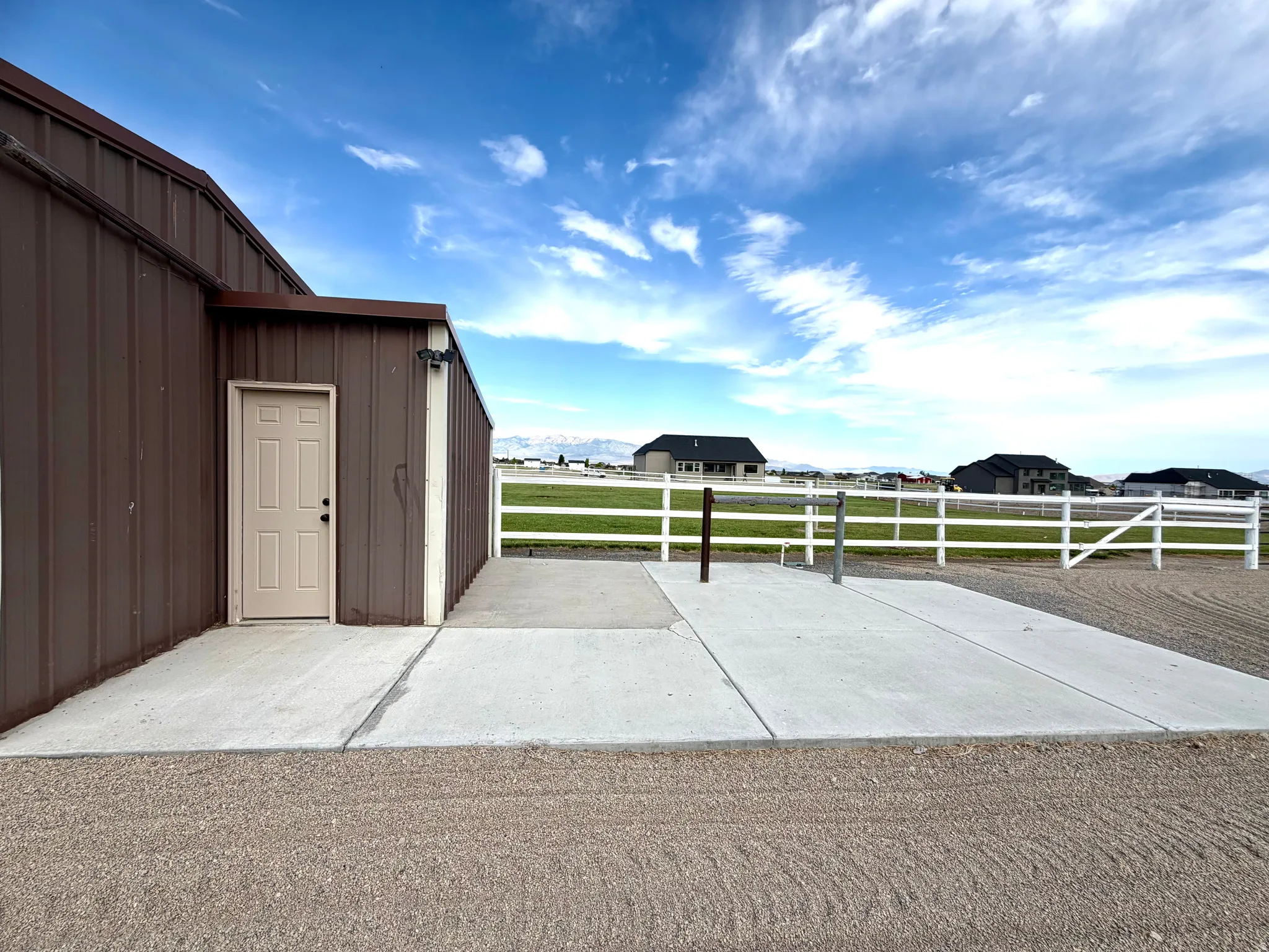 Brown corrugated metal building with a light-colored entry door, concrete pad, and white perimeter fencing