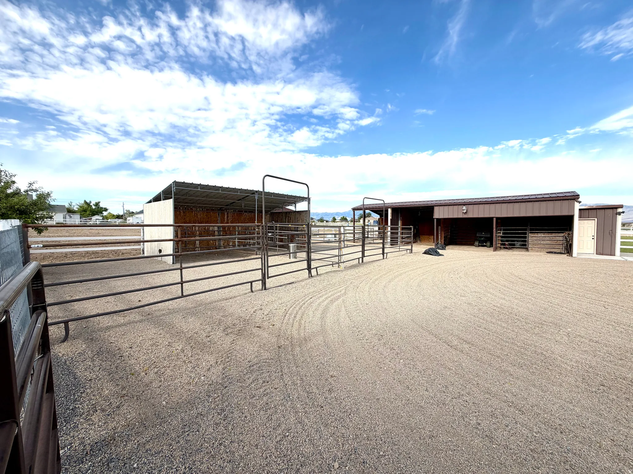 Expansive gravel-surfaced equestrian area featuring multiple fenced enclosures, a covered livestock shelter with a metal roof, and an additional barn structure with exterior stalls