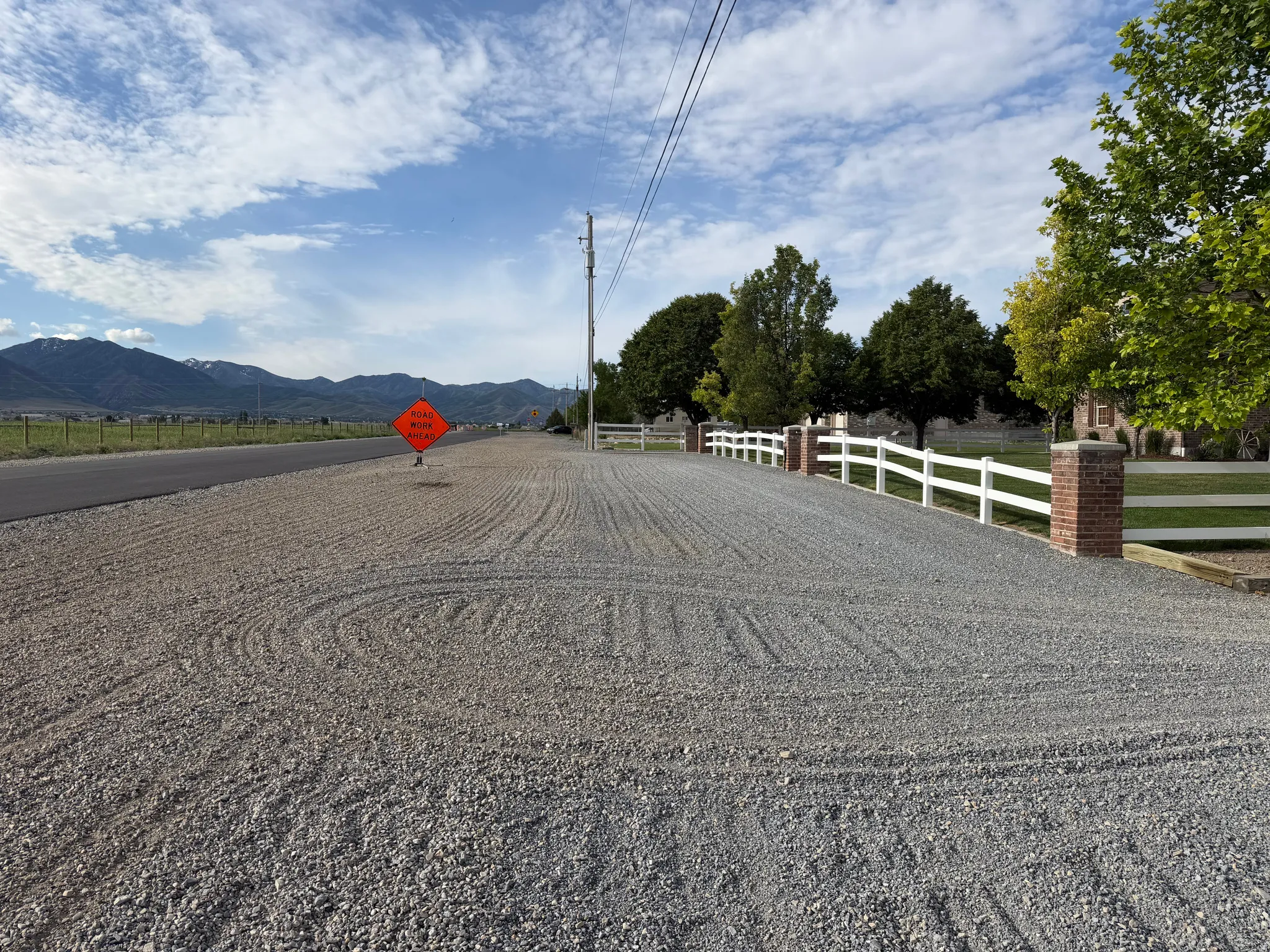 Gravel driveway flanked by a white fence with brick pillars, set against a backdrop of mature trees and distant mountains