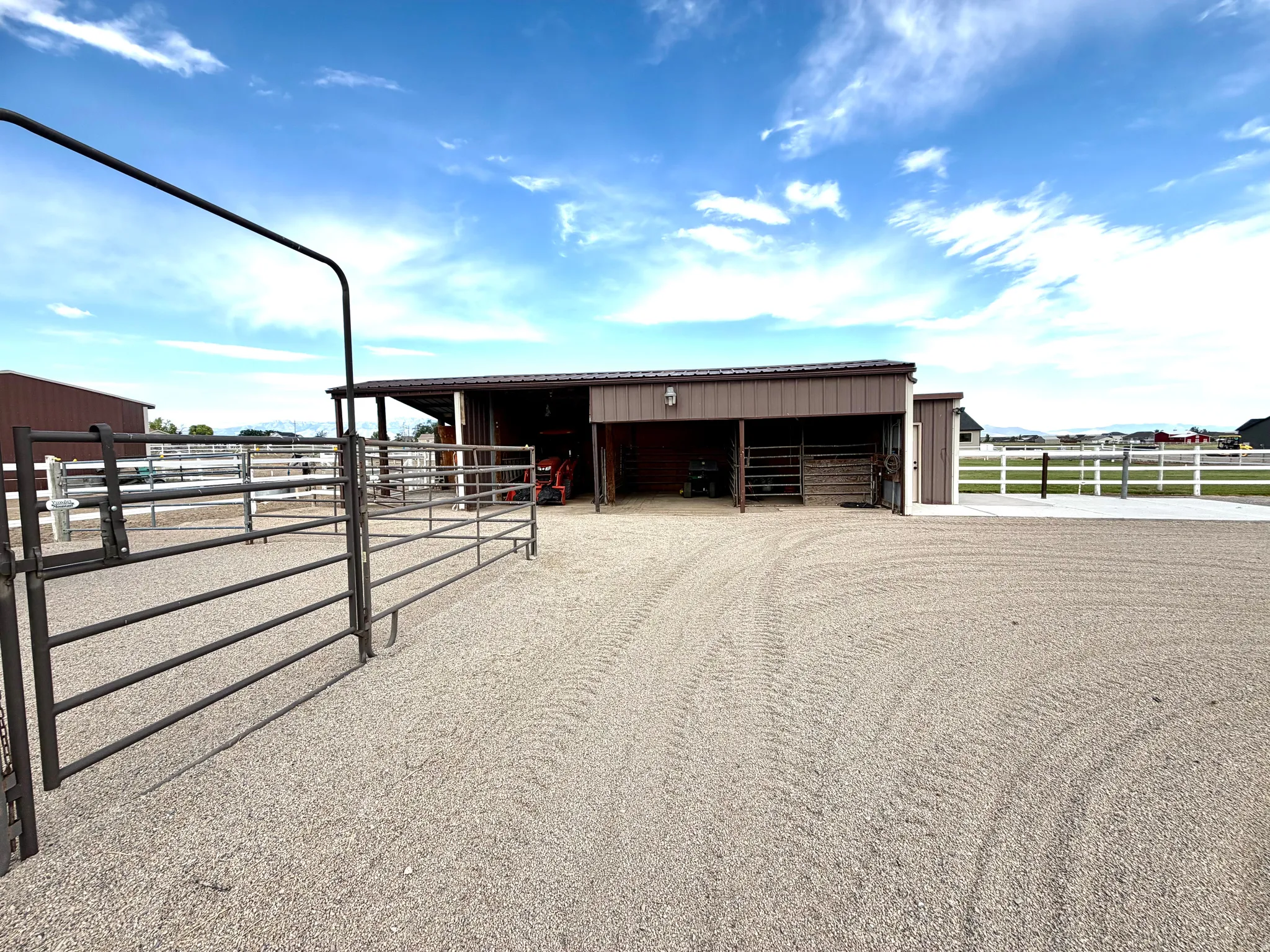 Gravel turnout area featuring metal fencing, a covered barn structure with multiple stalls, a white perimeter fence, and concrete paving