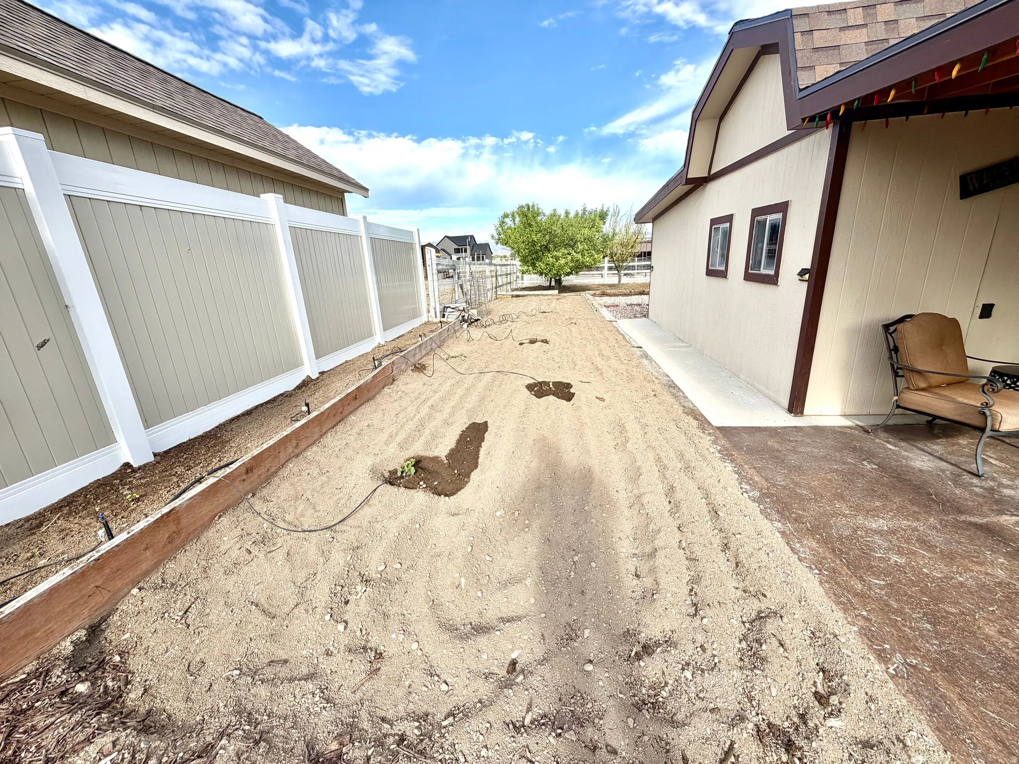 Side yard featuring a cultivated garden bed with drip irrigation, a light-colored privacy fence, and a concrete pathway leading to a building with a brown shingle roof