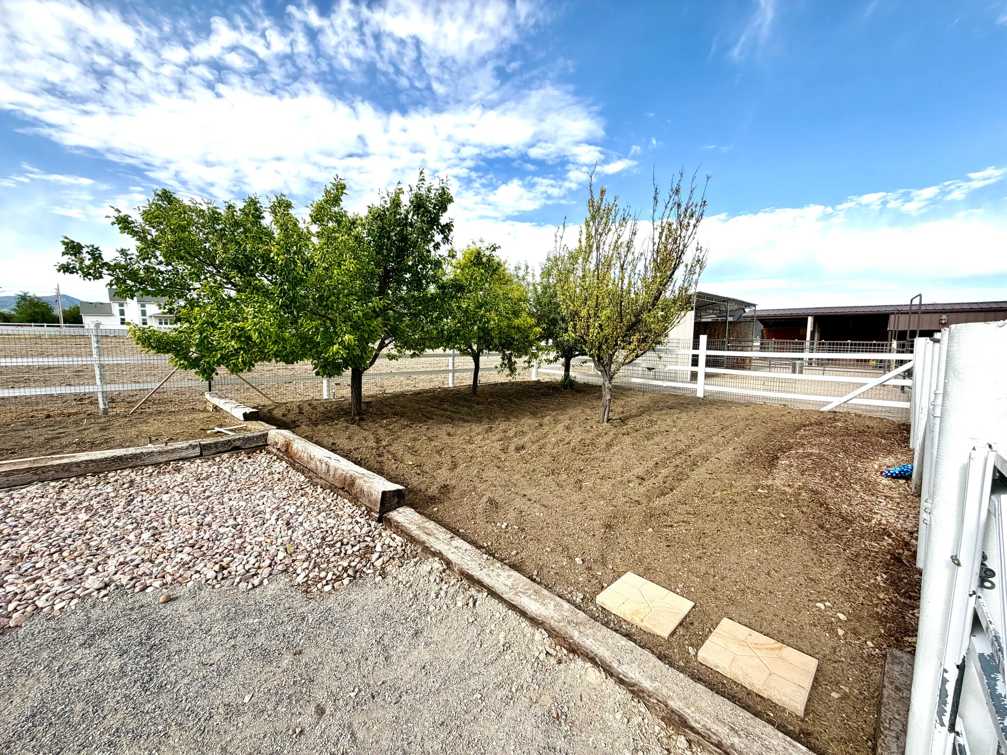 Outdoor area featuring established trees, a white perimeter fence, and a gravel surface