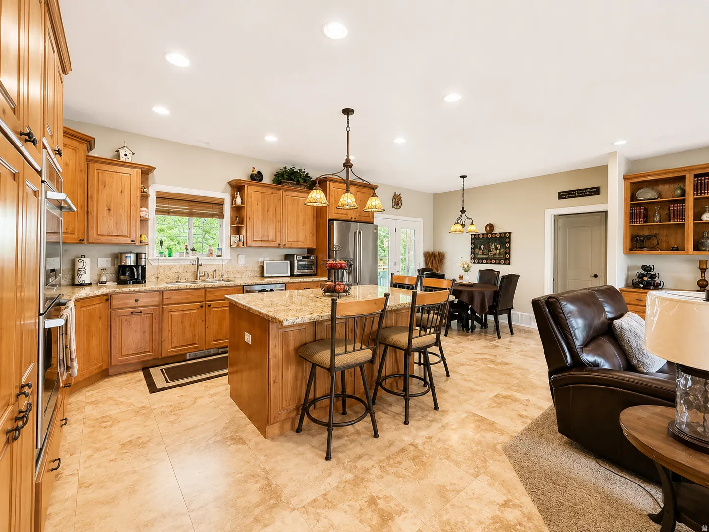 Open-concept kitchen featuring light wood cabinetry, stone-finish countertops, stainless steel appliances, and tile flooring