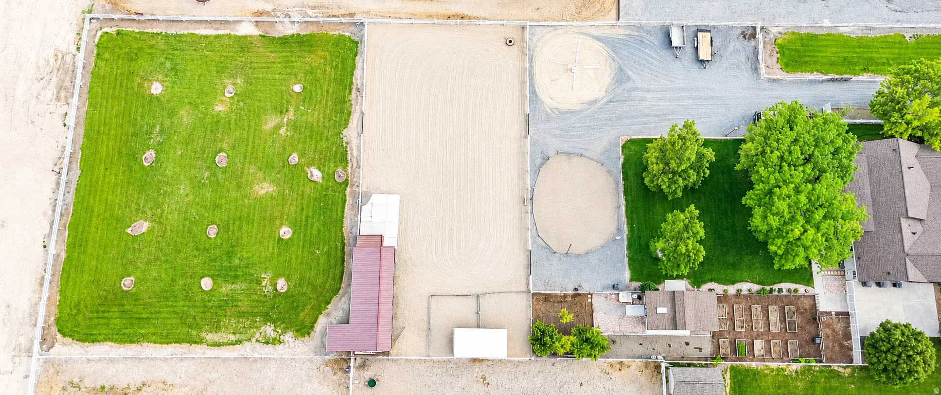 Aerial view showcasing extensive grounds with a large green pasture, an expansive sand arena, a barn structure with a reddish-brown roof, a gravel drive, and mature trees providing shade