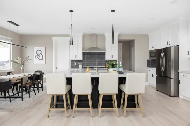 Open-concept kitchen featuring a large central island with seating, stainless steel appliances, white cabinetry with dark hardware, a dark subway tile backsplash, and light wood-finish flooring